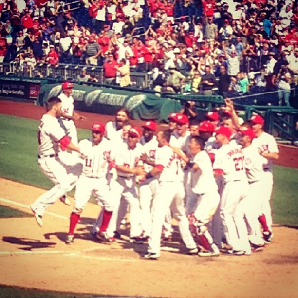 Baseball players in white and red uniforms celebrate together on the field while fans cheer in the crowded stadium stands behind them.