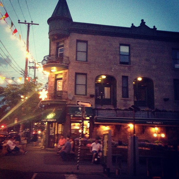 A brick building with a distinctive turret corner houses a busy restaurant or bar with outdoor seating, where many people are dining as evening approaches with warm lighting illuminating the scene.
