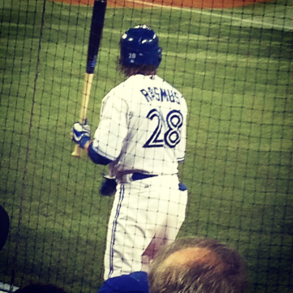 A baseball player wearing jersey number 28 with "RASMUS" on the back stands at home plate holding a bat, viewed from behind through protective netting.