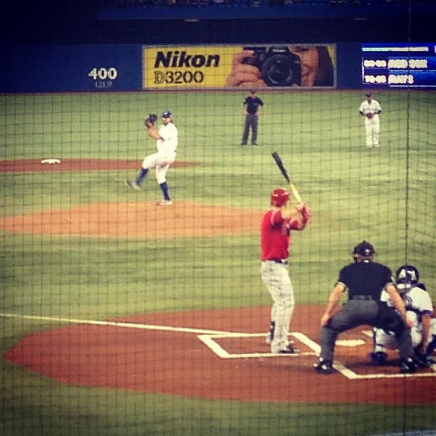 A baseball game in progress showing a batter in red uniform at home plate, with the catcher, umpire, and fielders positioned on the field under stadium lights.