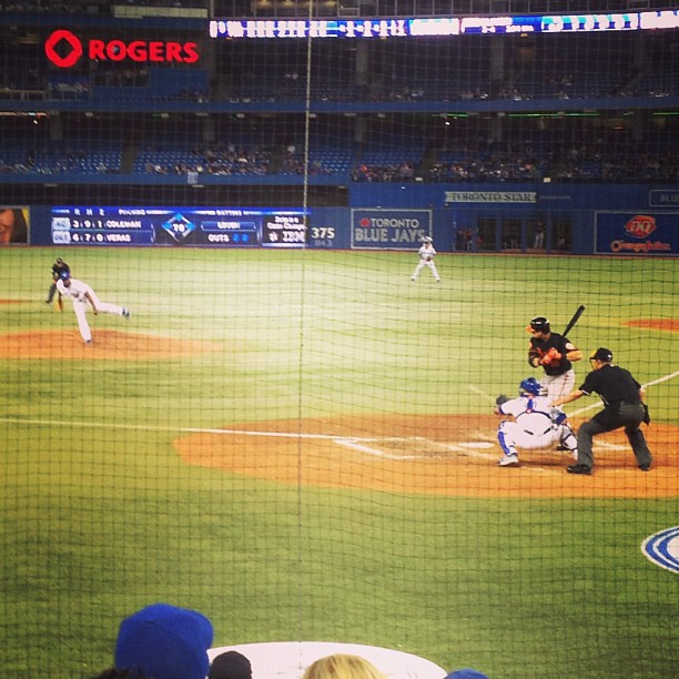 A baseball game in progress at Rogers Centre showing a batter at home plate with catcher and umpire, while a pitcher delivers from the mound and fielders are positioned around the diamond.