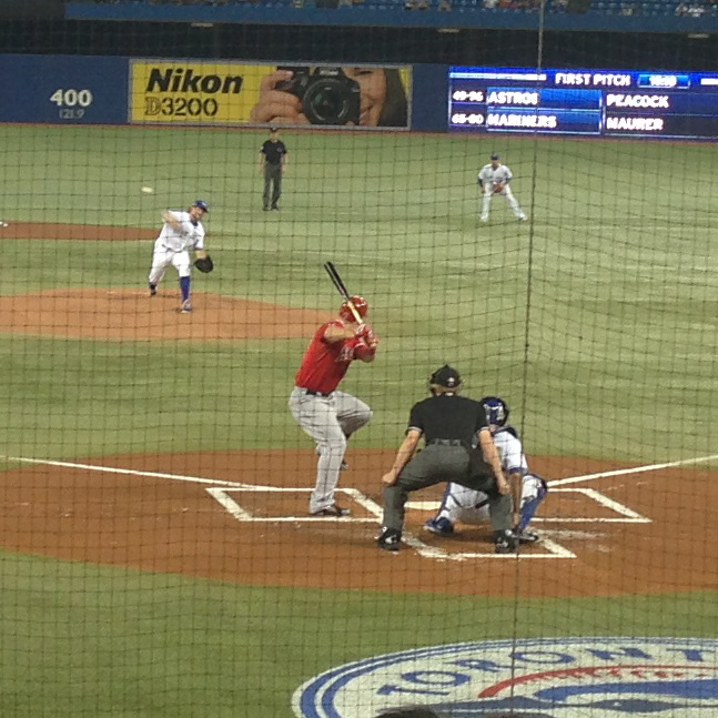 A baseball game in progress showing a batter in red uniform at home plate, with a catcher, umpire, and pitcher visible on the field, along with stadium advertising boards in the background.