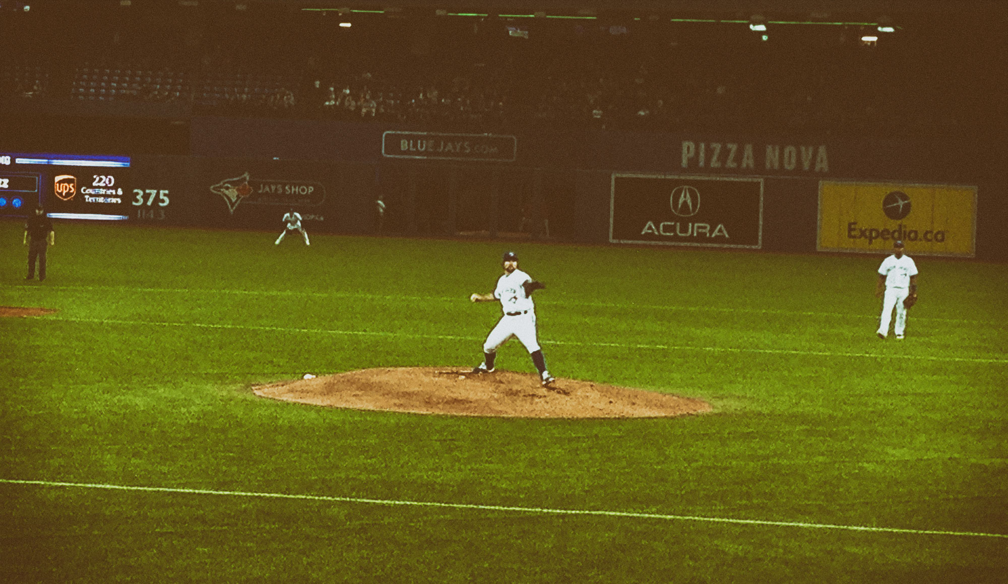 A baseball pitcher in mid-throw on the mound during a nighttime game, with outfield advertising boards visible in the background including sponsors like Pizza Nova, Acura, and Expedia.