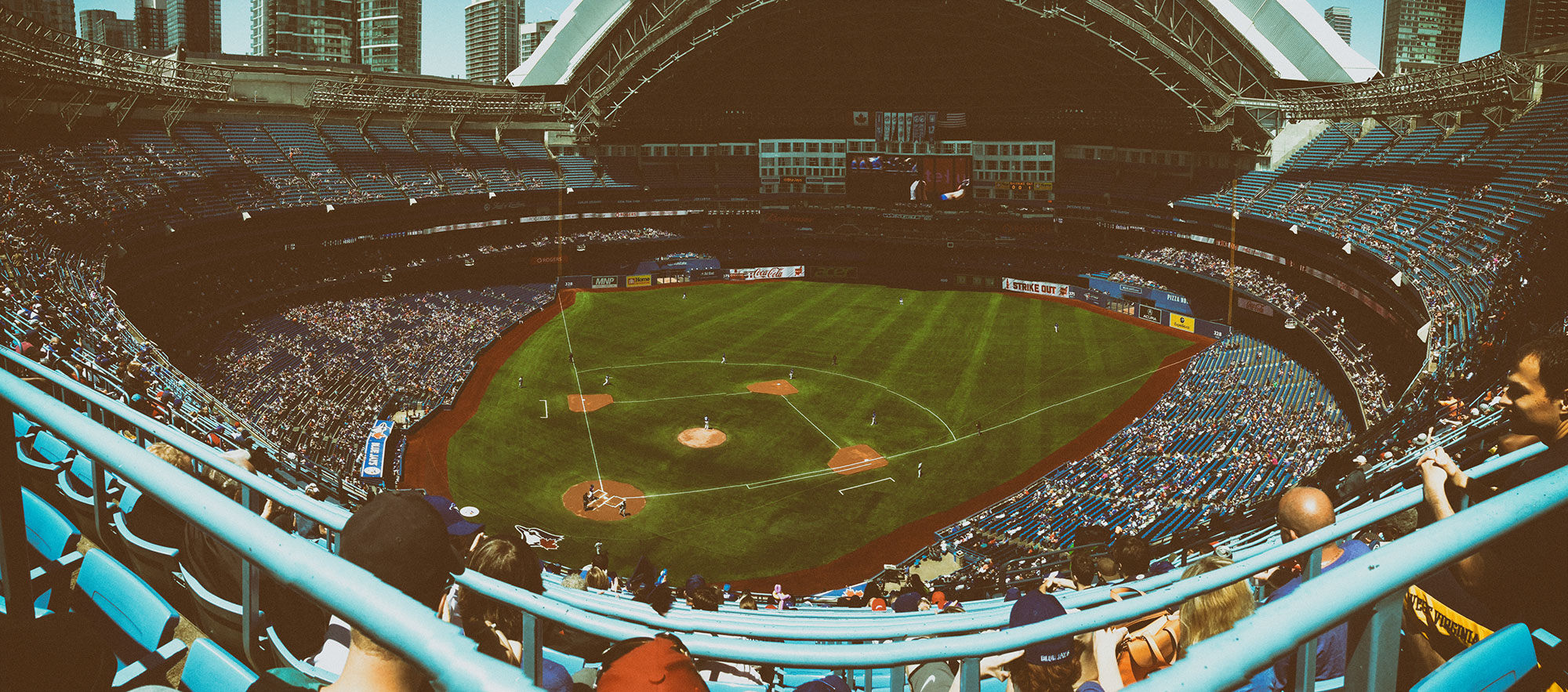 A panoramic view of a baseball stadium from the upper deck showing the full diamond-shaped field with players positioned during a game, surrounded by tiered seating filled with spectators and city skyscrapers visible in the background.