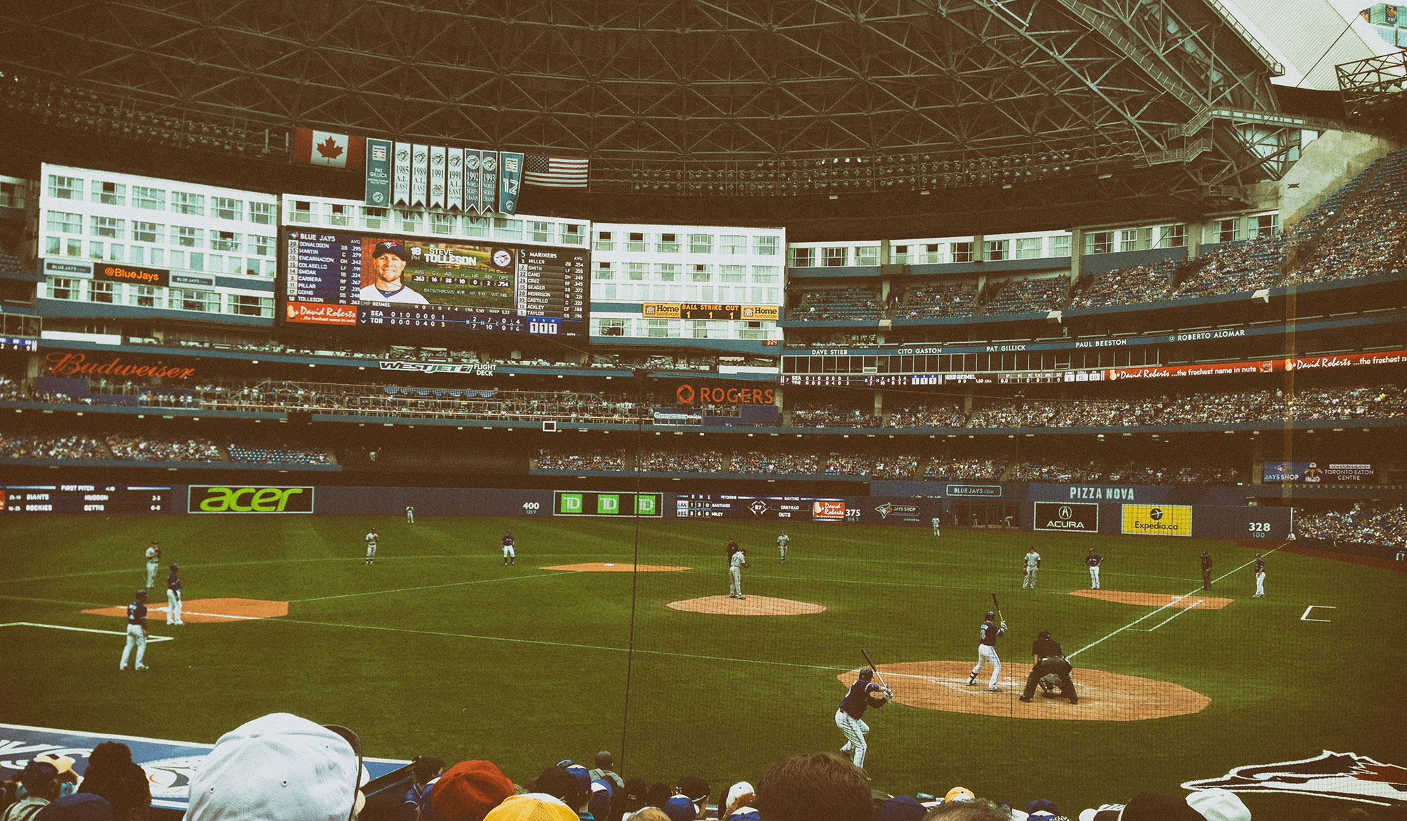 A baseball game in progress at Rogers Centre stadium in Toronto, with players on the field, crowded stands, and various advertising displays and scoreboards visible around the dome-shaped venue.