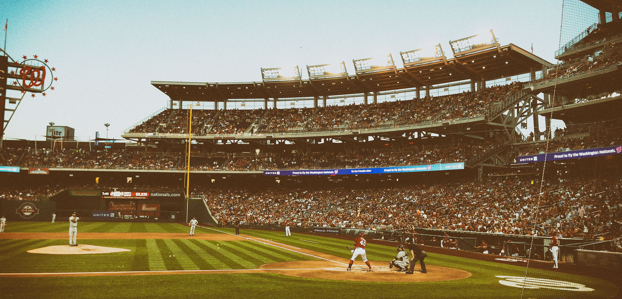 A baseball game in progress at a large stadium with packed stands, showing players positioned on the field including a batter at home plate, with the distinctive multi-tiered architecture and lighting visible under a clear sky.