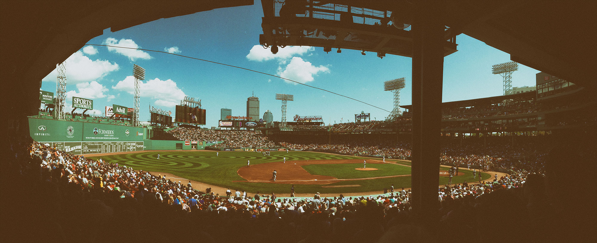 A view of a baseball game at Fenway Park taken from under the stadium's overhang, showing the iconic Green Monster wall, filled stands, city skyline, and players on the field during daytime.