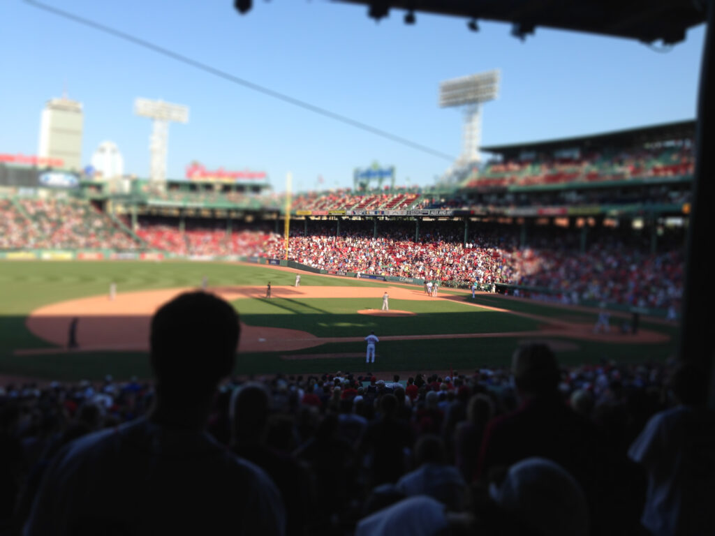 A baseball game in progress at Fenway Park, showing the iconic green outfield wall and packed stadium seating filled with fans under a clear blue sky.