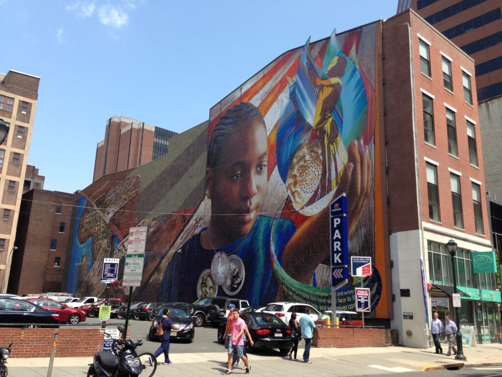 A large colorful mural covers the side of a brick building, featuring a portrait of a person surrounded by vibrant geometric patterns and symbolic imagery, with a parking lot and urban street scene in the foreground.