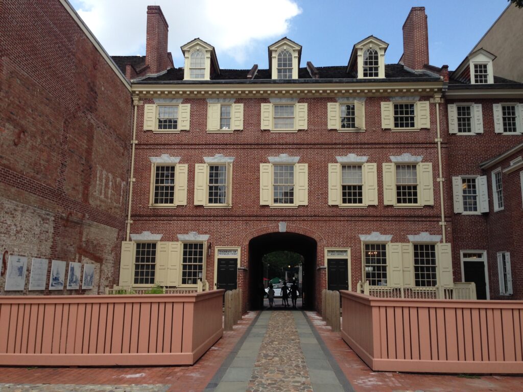 A historic red brick colonial building with cream-colored shutters and dormers, featuring a central arched passageway entrance accessed by a wooden walkway over cobblestones.