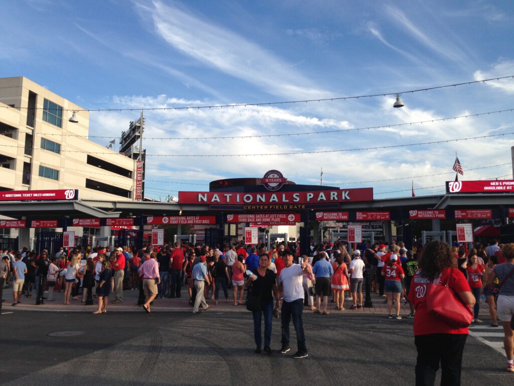 Crowds of baseball fans wearing red gather outside the Nationals Park centerfield gate entrance under a blue sky with wispy clouds.