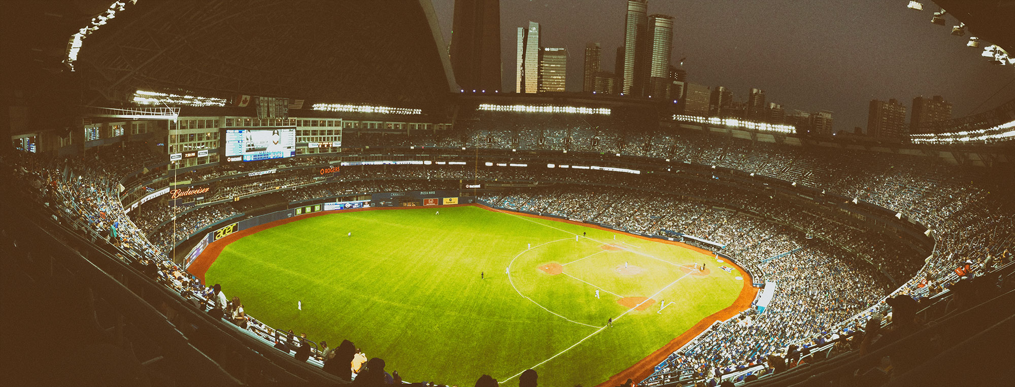 A panoramic view of a large baseball stadium at night with bright green field lighting, filled stands of spectators, and a city skyline with tall buildings visible in the background.