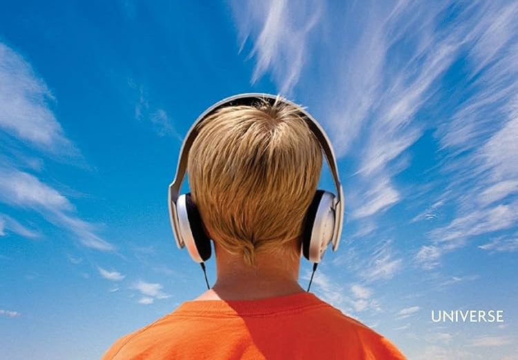 A person in an orange shirt seen from behind, wearing large headphones and looking up at a bright blue sky with wispy clouds.