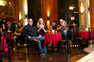A group of people laughing and enjoying a show at a dimly lit club with ornate, Gothic-style interiors and red tablecloths on the tables.