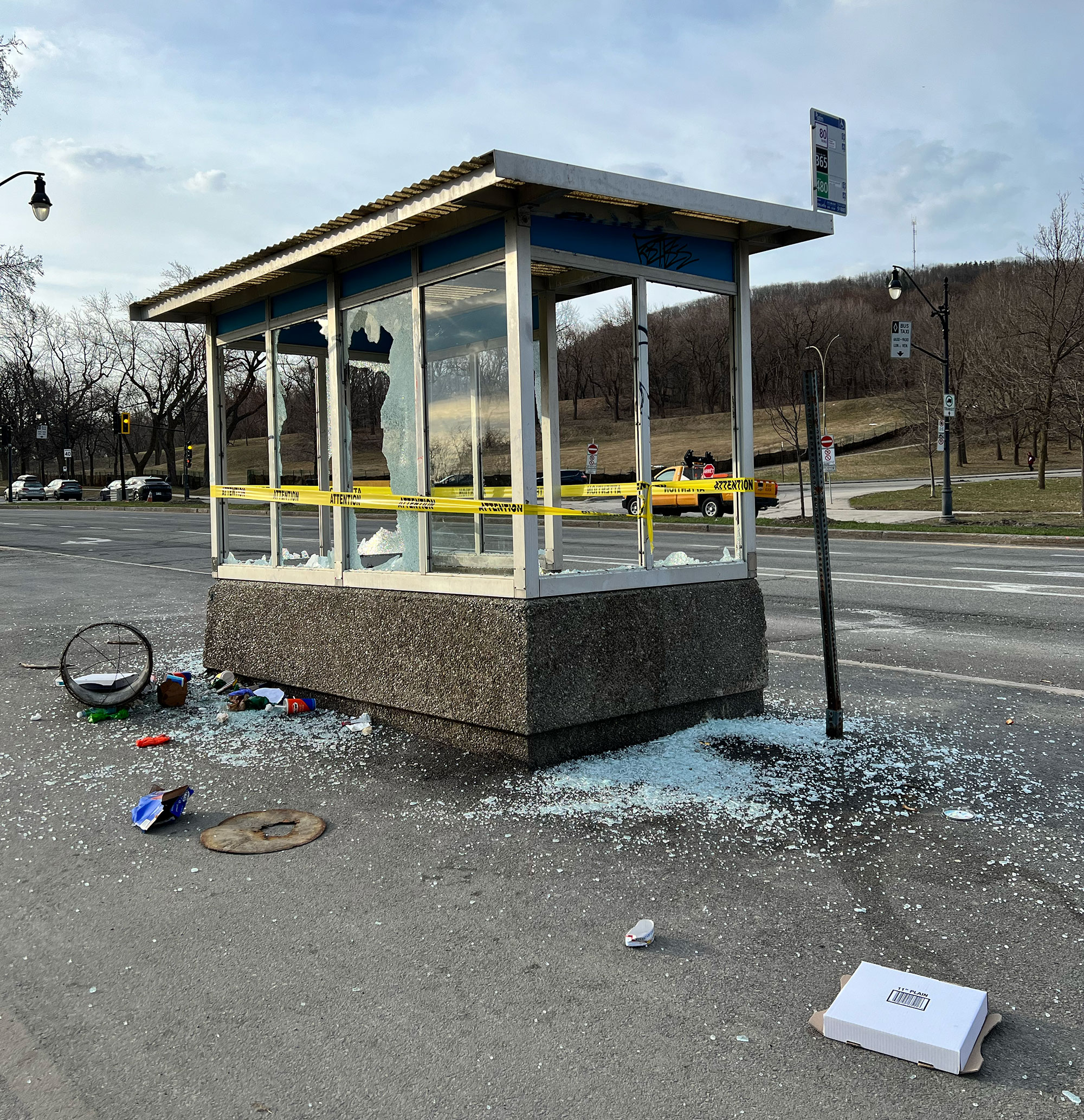 Damaged bus shelter with shattered glass and yellow caution tape, surrounded by debris including a bicycle wheel and a pizza box, with a road and park in the background.