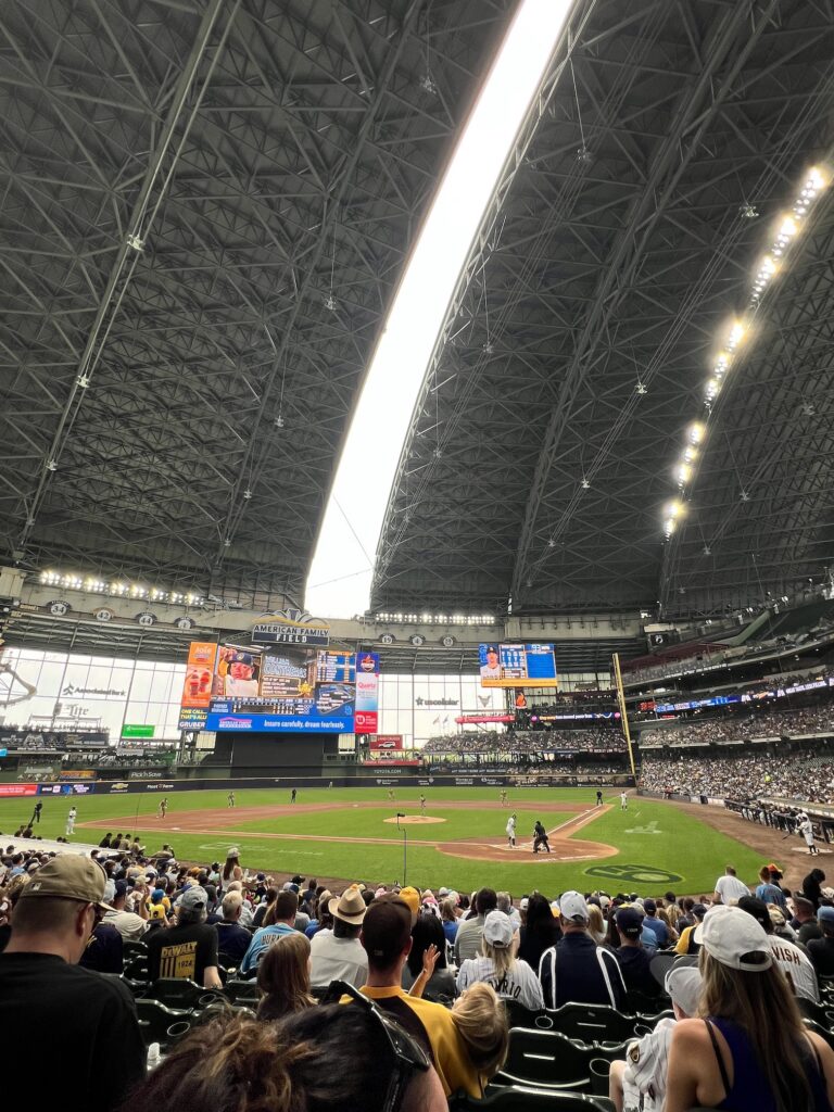 A baseball game in progress at a large indoor stadium with a retractable roof partially open, showing players on the field and crowds of spectators in the stands under bright stadium lighting.