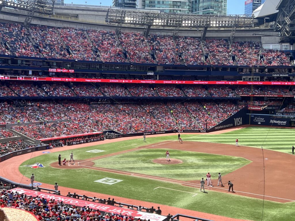 A baseball game in progress at a packed stadium with red seats filled with spectators, showing players positioned on the field during play with city buildings visible beyond the stadium walls.