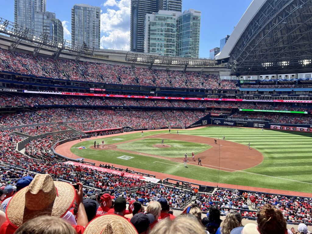 A packed baseball stadium during a game, with thousands of spectators in the stands watching players on the field, surrounded by tall city buildings under a blue sky with white clouds.