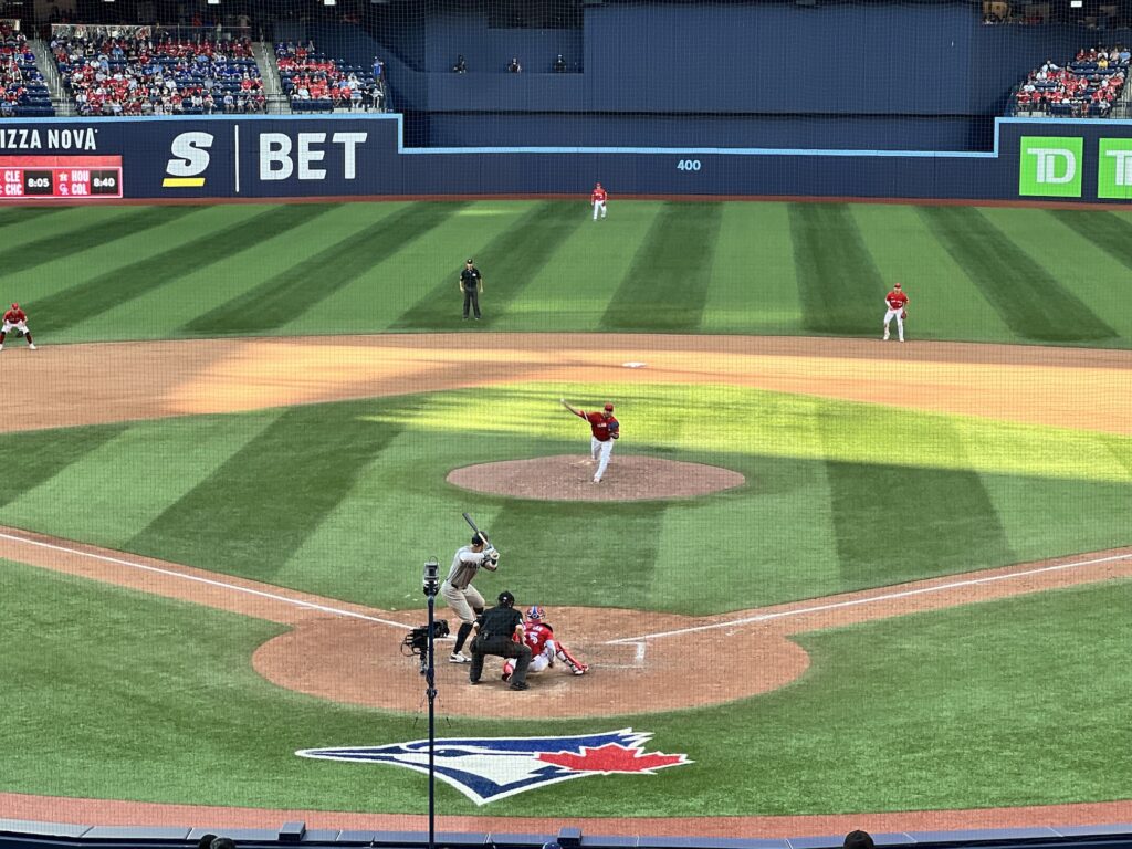 A baseball game in progress at a stadium with a pitcher on the mound throwing to a batter at home plate, with fielders positioned around the diamond and the Toronto Blue Jays logo visible behind home plate.