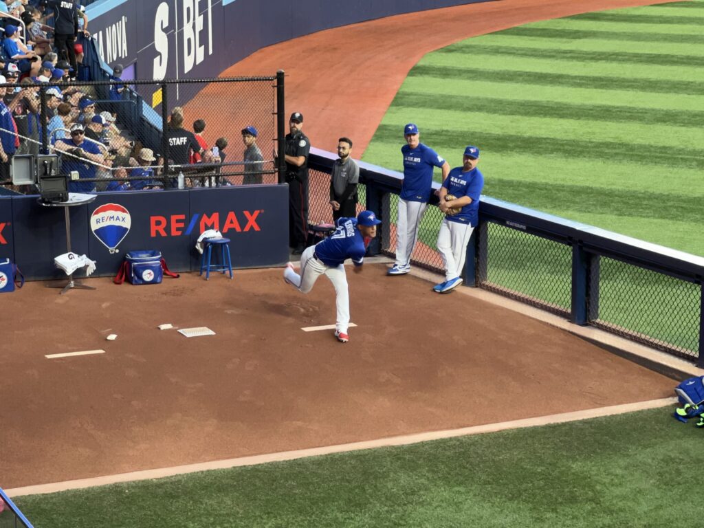 A baseball player in a blue uniform practices pitching in the bullpen area while teammates and staff watch from behind a chain-link fence, with stadium seating visible in the background.