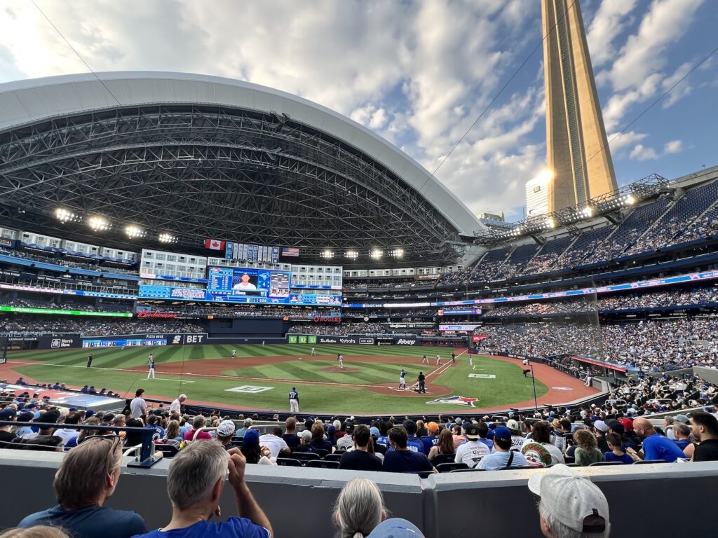 A baseball game in progress at a large domed stadium with spectators filling the blue seats and the CN Tower visible in the background. Players are positioned on the field during gameplay under the stadium's distinctive curved roof structure.