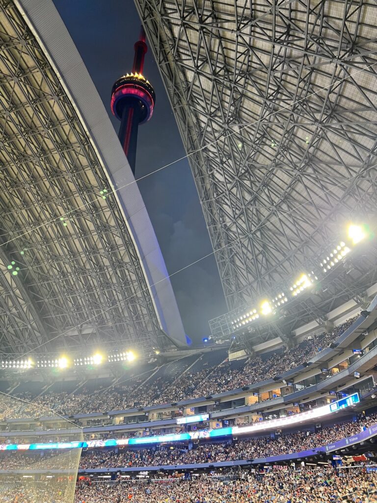 Interior view of a crowded stadium with a retractable roof opening, showing the CN Tower illuminated with red lights visible through the opening against the evening sky.