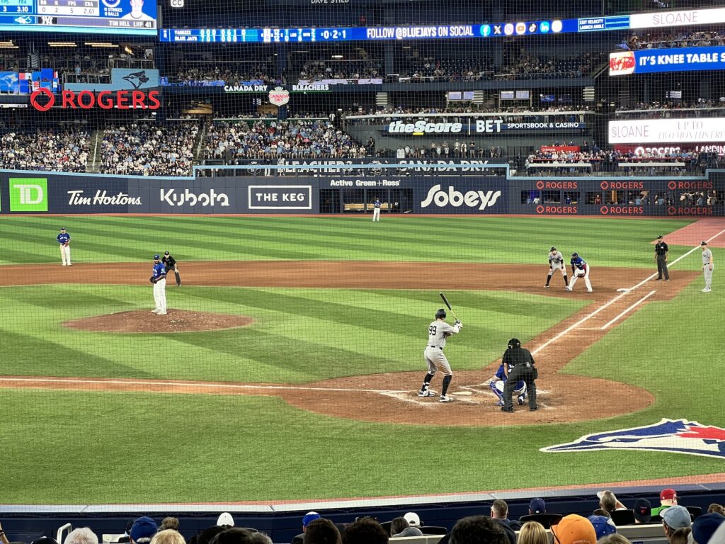 A baseball game in progress at Rogers Centre in Toronto, with a batter at home plate, pitcher on the mound, and fielders positioned around the diamond while spectators fill the stadium seats.