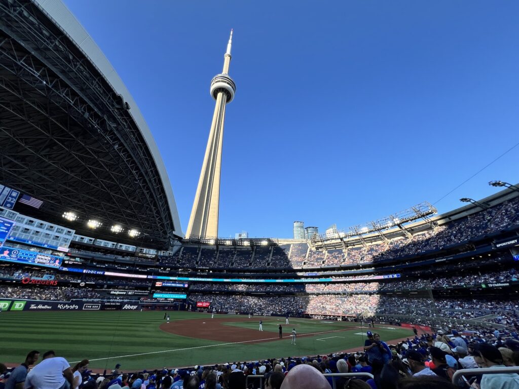 A baseball stadium filled with spectators during a game, with Toronto's CN Tower prominently visible rising above the stadium against a clear blue sky.