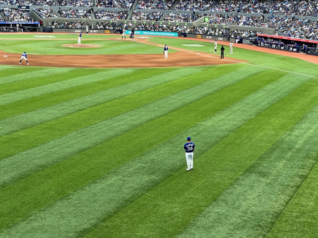 A baseball game in progress at a large stadium, with a player wearing jersey number 30 standing in the outfield on pristinely manicured grass with distinctive mowing patterns, while other players are positioned around the infield and packed stands are visible in the background.