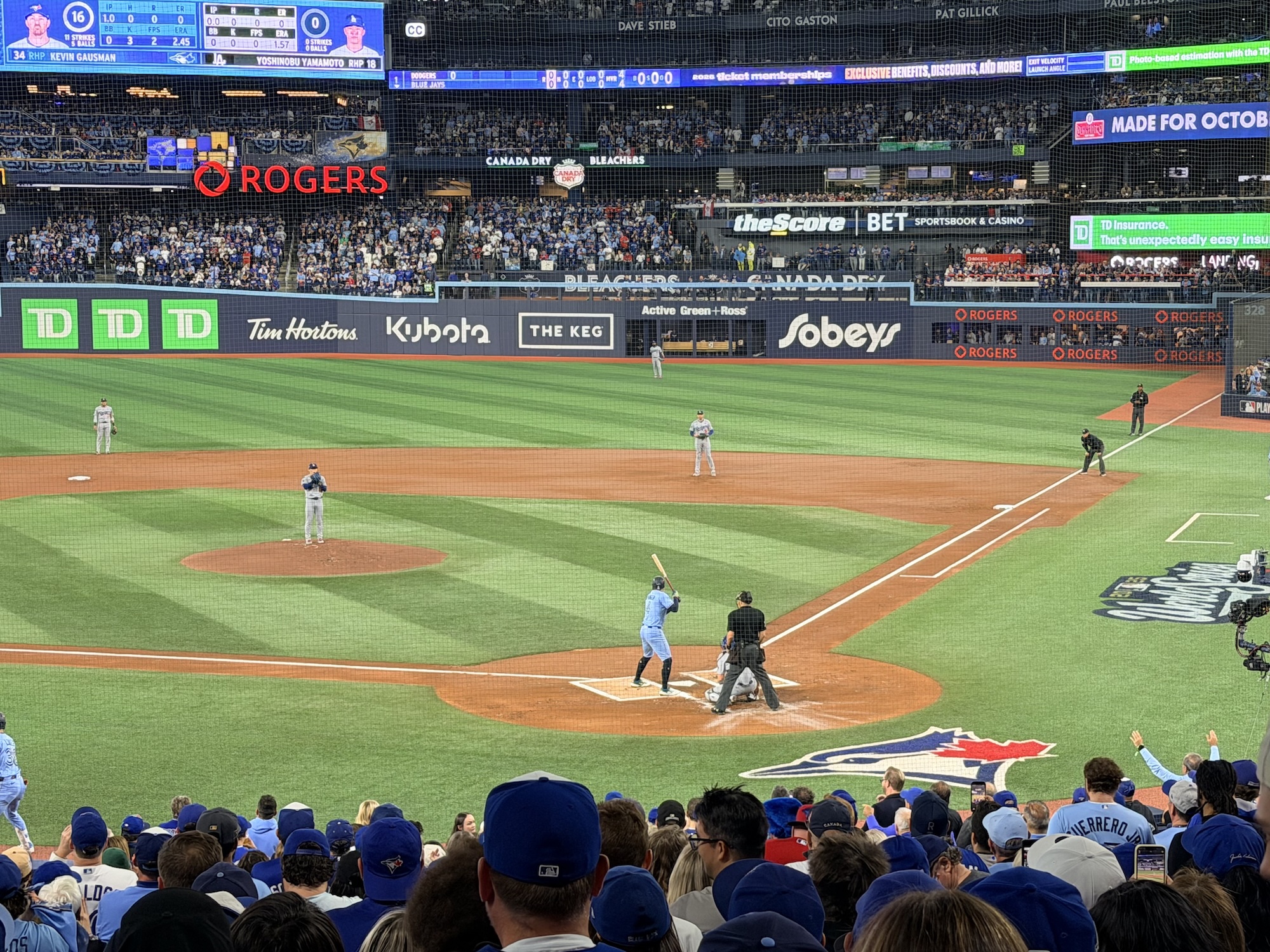 A baseball game in progress at Rogers Centre with a batter in light blue uniform at home plate, fielders positioned around the diamond, and crowds of fans wearing predominantly blue team colors filling the stadium seats.