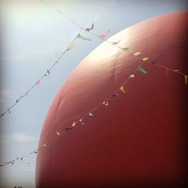 Colorful prayer flags on strings stretch across a red rock formation against a bright sky with sunlight creating lens flare effects.
