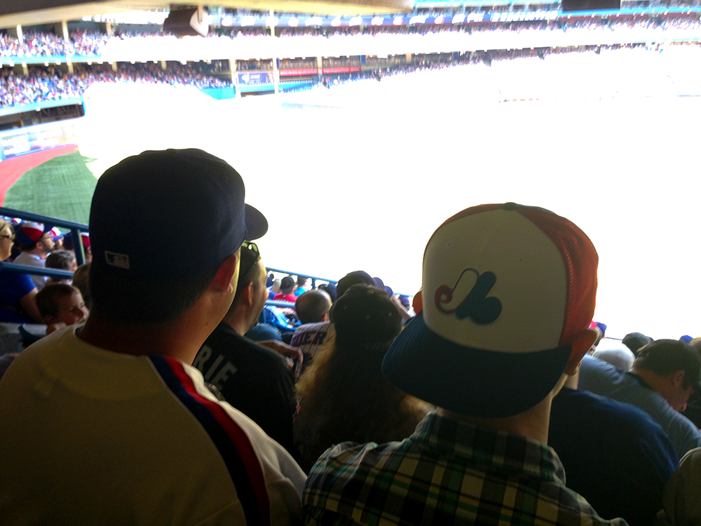 Two baseball fans in caps sit in stadium seats overlooking a bright, overexposed baseball field with visible stands and spectators in the background.