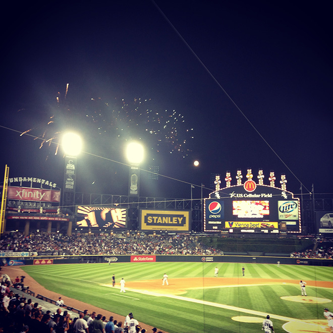 A nighttime baseball game at a stadium with fireworks exploding in the sky above the field, illuminating the crowded stands and large outfield scoreboards displaying sponsor logos.