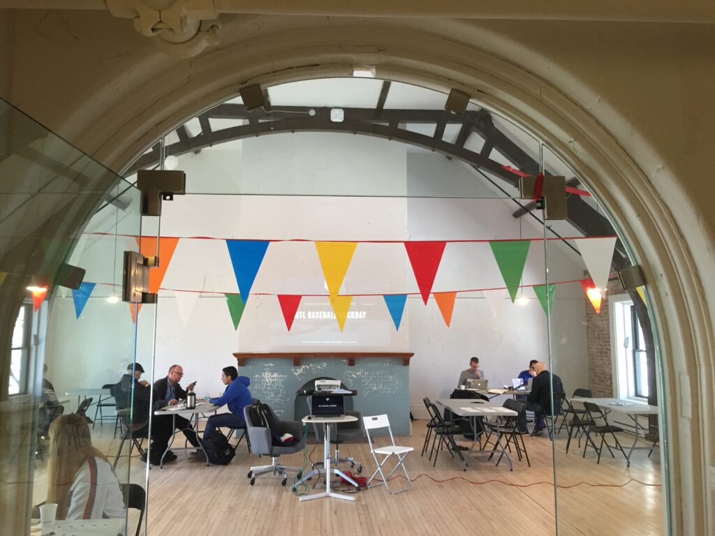 A classroom or meeting space with arched doorway framing the view, featuring colorful triangle bunting hanging across white walls, several people seated at tables working, and a chalkboard visible in the background.