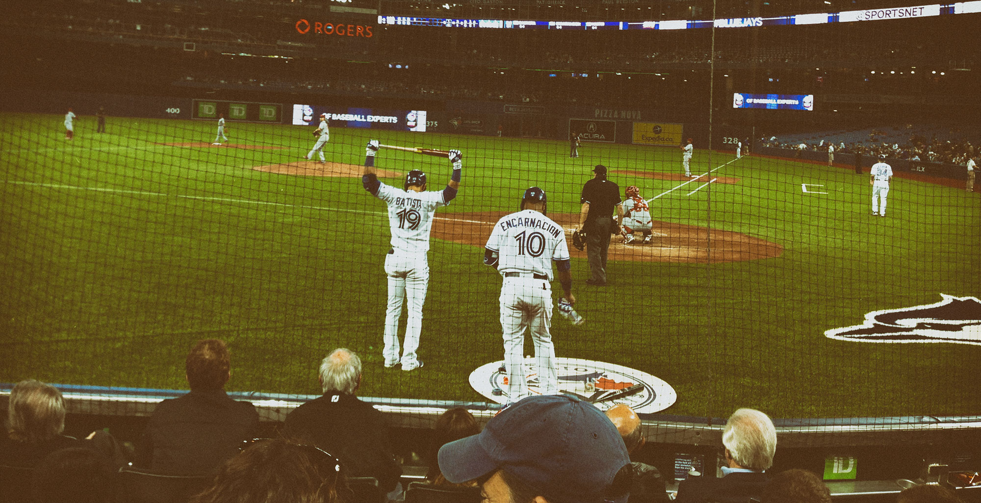 A baseball player wearing jersey number 19 celebrates with arms raised at home plate while teammate number 10 (Encarnacion) approaches, with spectators visible in the foreground at what appears to be Rogers Centre.
