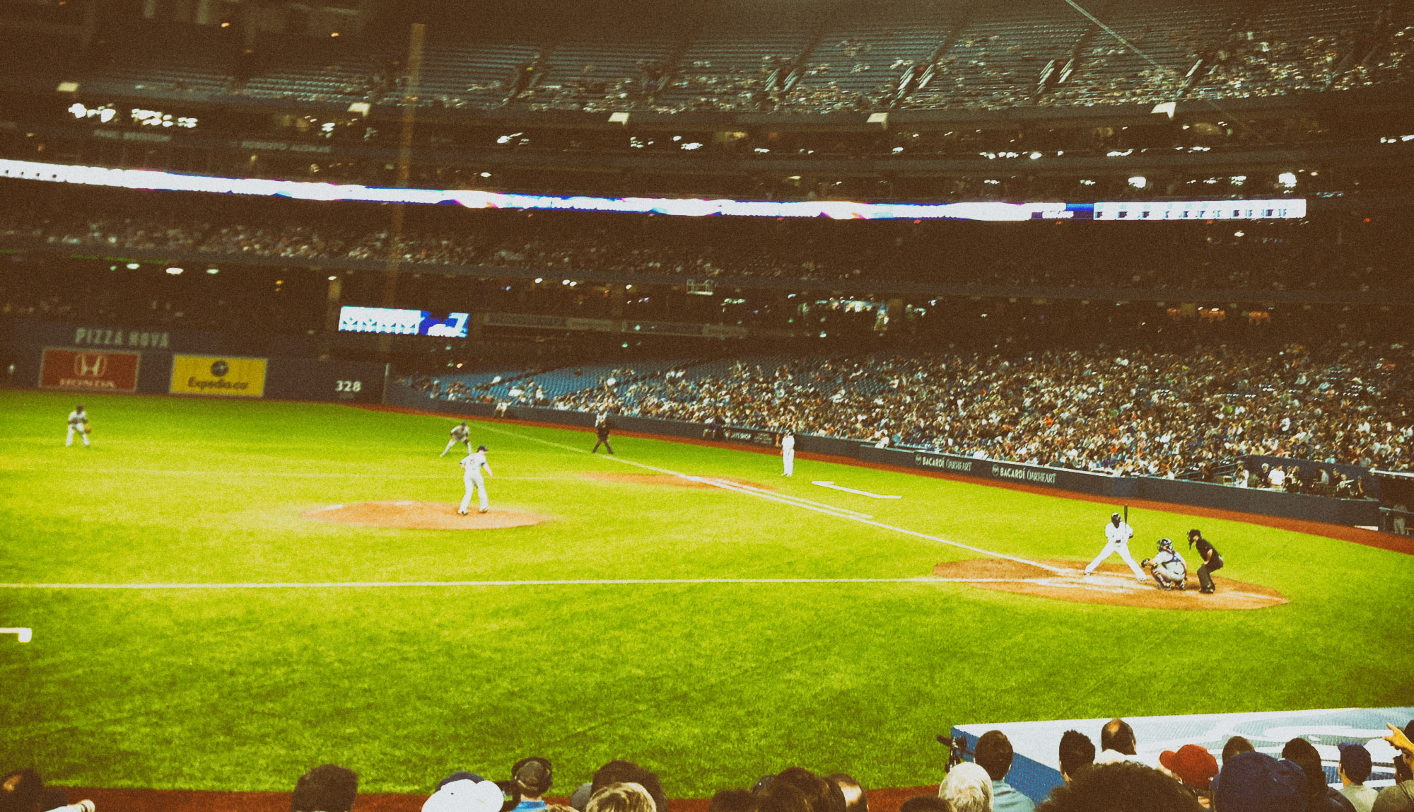 A baseball game in progress at a large stadium with players positioned on the bright green field and crowds filling the multi-tiered seating areas under artificial lighting.