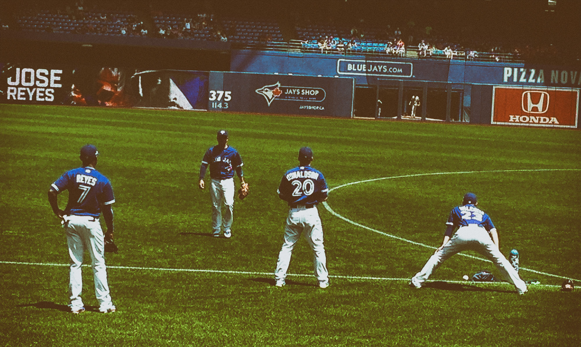 Four Toronto Blue Jays players in blue and white uniforms are positioned on a baseball field during a game, with one player crouched near first base while the others stand in the infield. The outfield wall displays various sponsor logos including Honda and references to Jose Reyes.