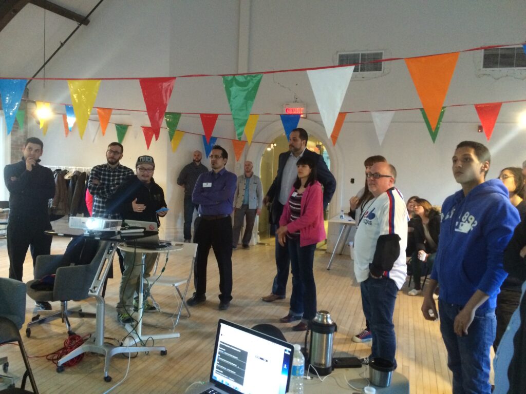 A group of people gathered in an office space with colorful triangular bunting hanging from the ceiling, watching a presentation or demonstration at a desk with computer equipment.