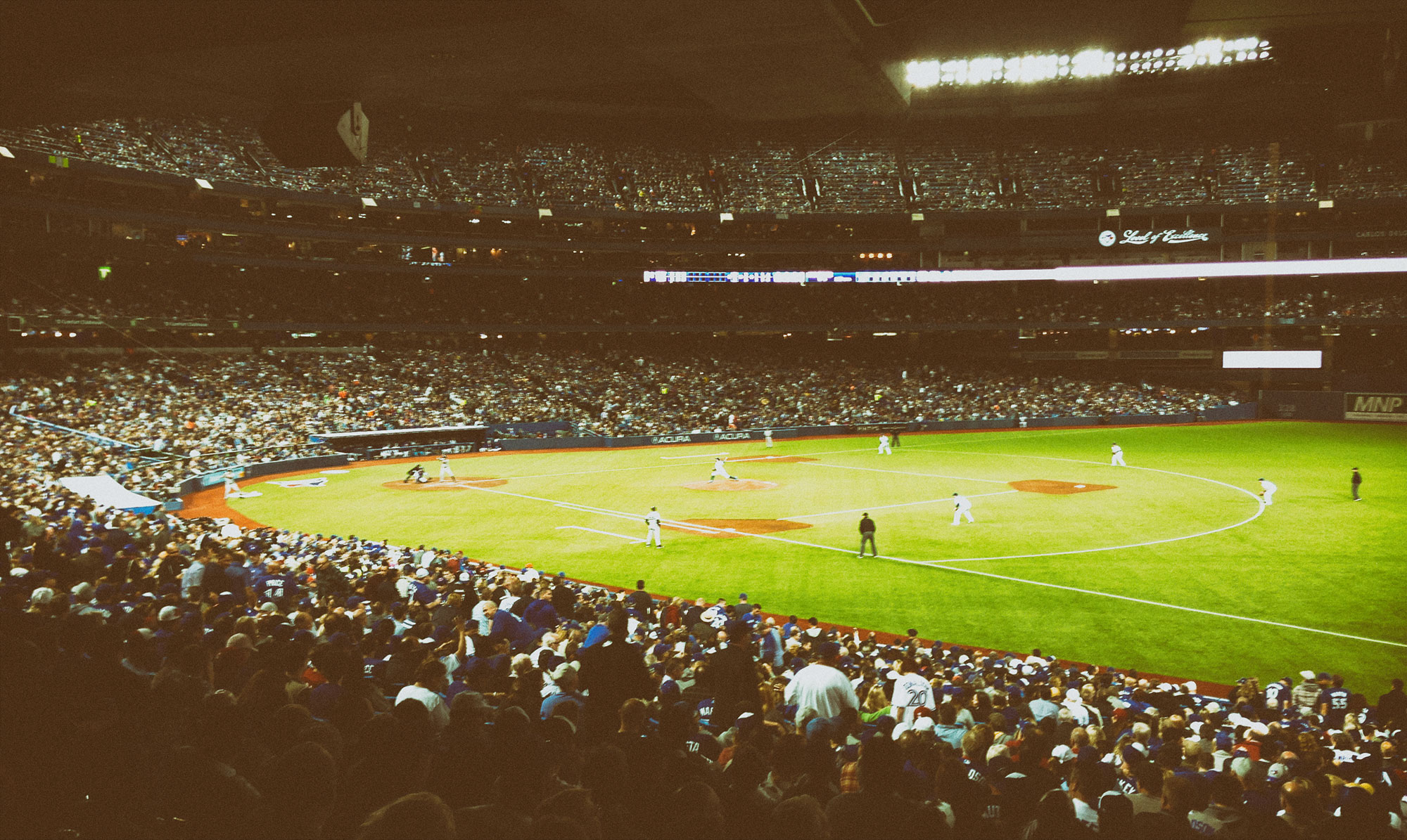 A packed baseball stadium at night with players positioned on the bright green field during a game, viewed from the stands with thousands of spectators filling the multi-tiered seating areas.