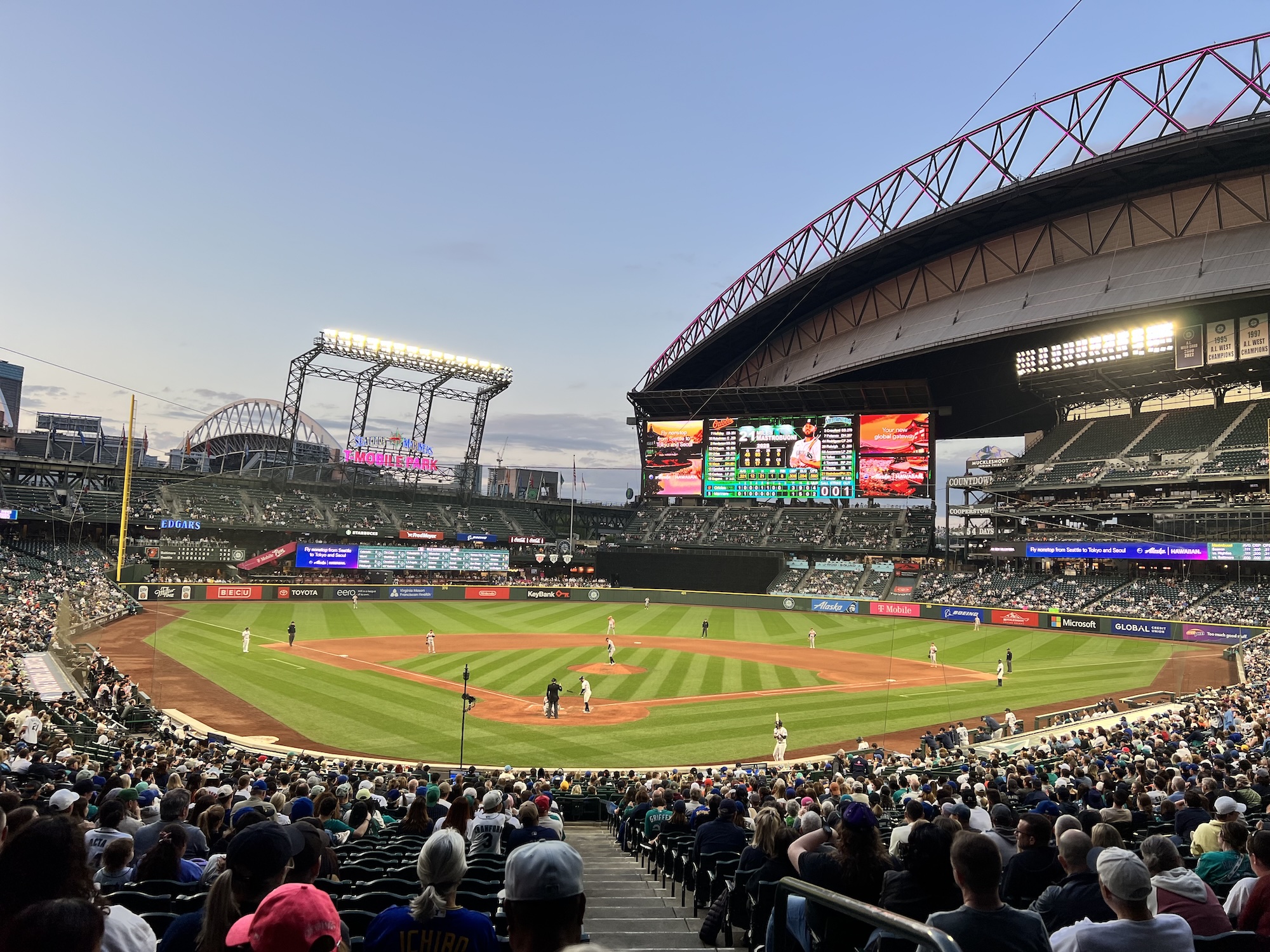 A baseball game in progress at a large stadium with crowds in the stands, featuring a retractable roof structure and large video scoreboard displaying game information under an evening sky.